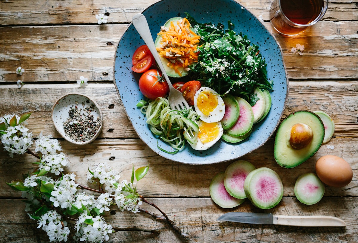 Colourful wellness bowl with avocado, soft-boiled egg, leafy greens, and radish on a rustic wooden table with spring blossoms